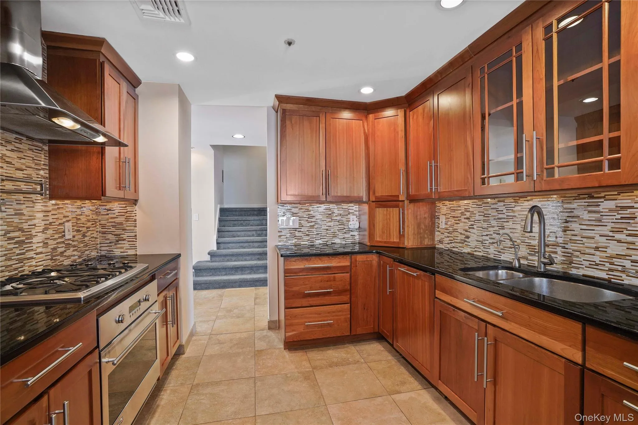 Kitchen with dark stone counters, wall chimney exhaust hood, brown cabinetry, stainless steel appliances, and glass insert cabinets Kitchen with dark stone counters, wall chimney exhaust hood, brown cabinetry, stainless steel appliances, and glass insert cabinets