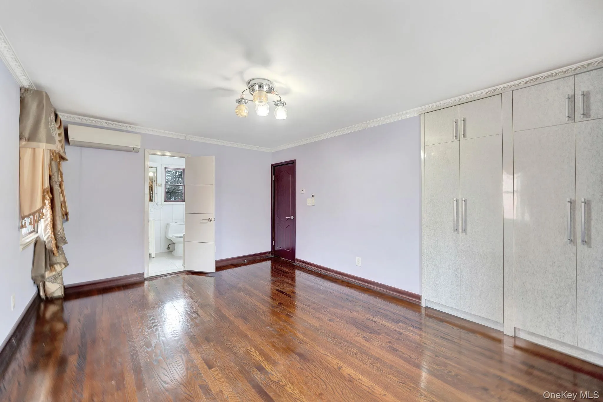 Unfurnished bedroom with a wall mounted air conditioner, connected bathroom, dark wood-style floors, and ornamental molding Unfurnished bedroom with a wall mounted air conditioner, connected bathroom, dark wood-style floors, and ornamental molding