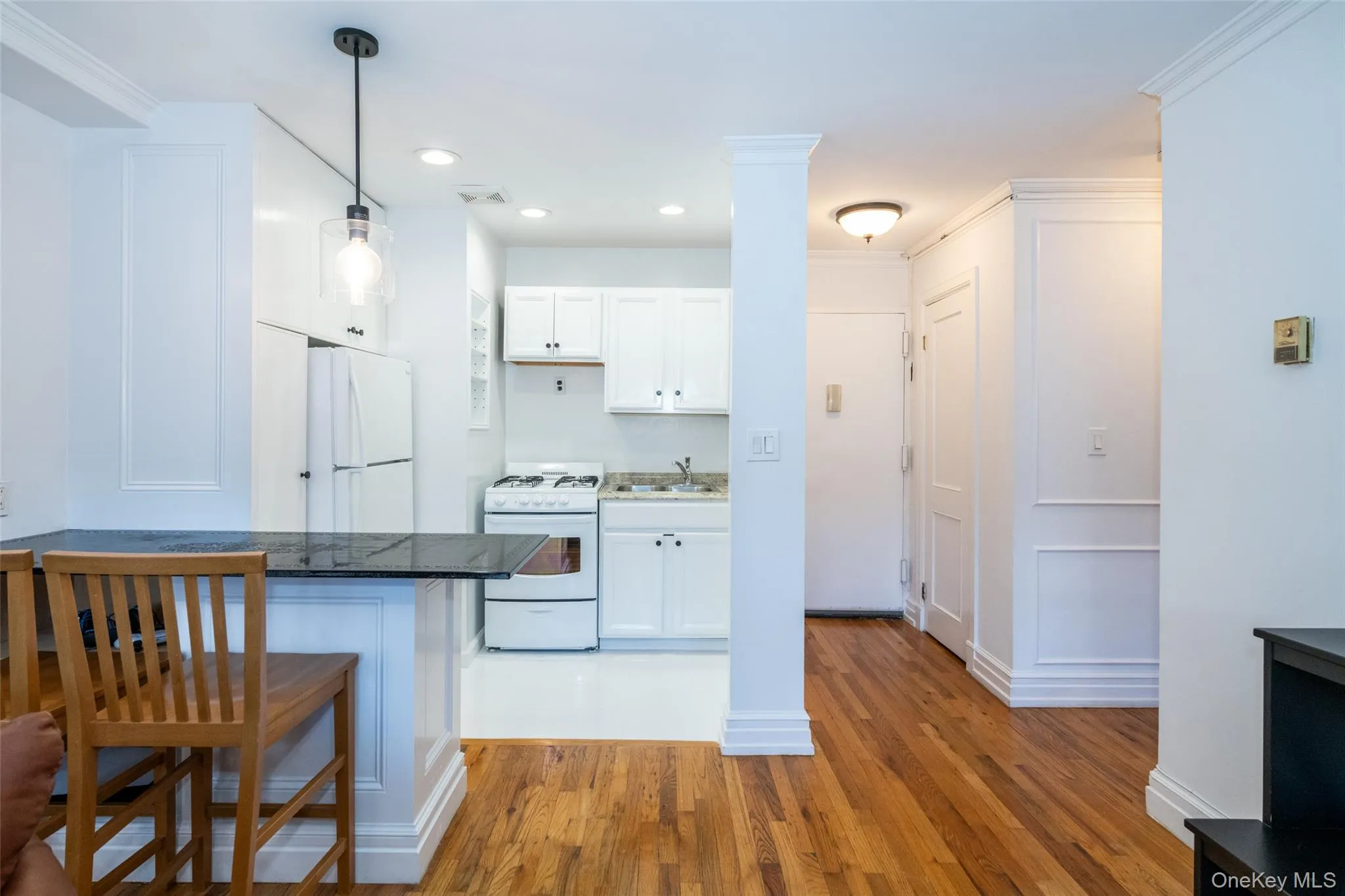 Kitchen featuring white cabinetry, white appliances, dark stone counters, light wood-type flooring, and hanging light fixtures Kitchen featuring white cabinetry, white appliances, dark stone counters, light wood-type flooring, and hanging light fixtures