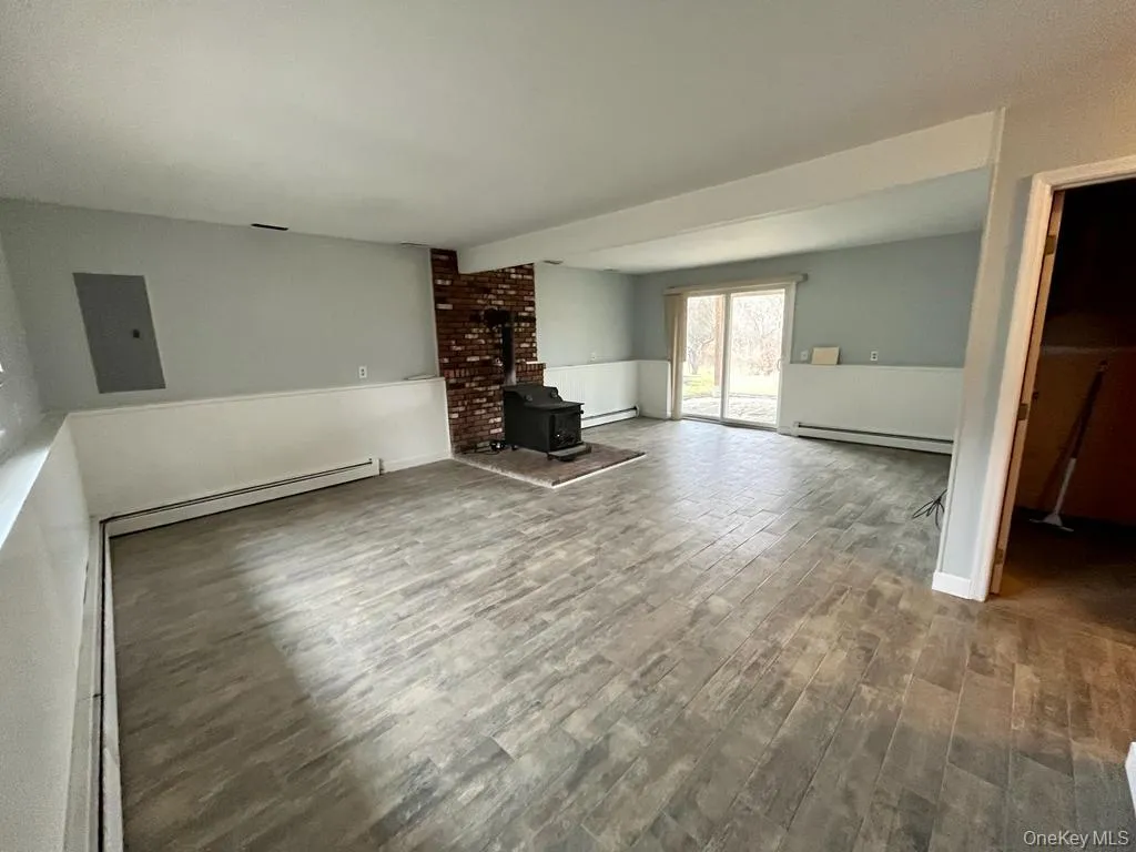 Unfurnished living room featuring a wood stove, electric panel, wood finished floors, and baseboard heating Unfurnished living room featuring a wood stove, electric panel, wood finished floors, and baseboard heating