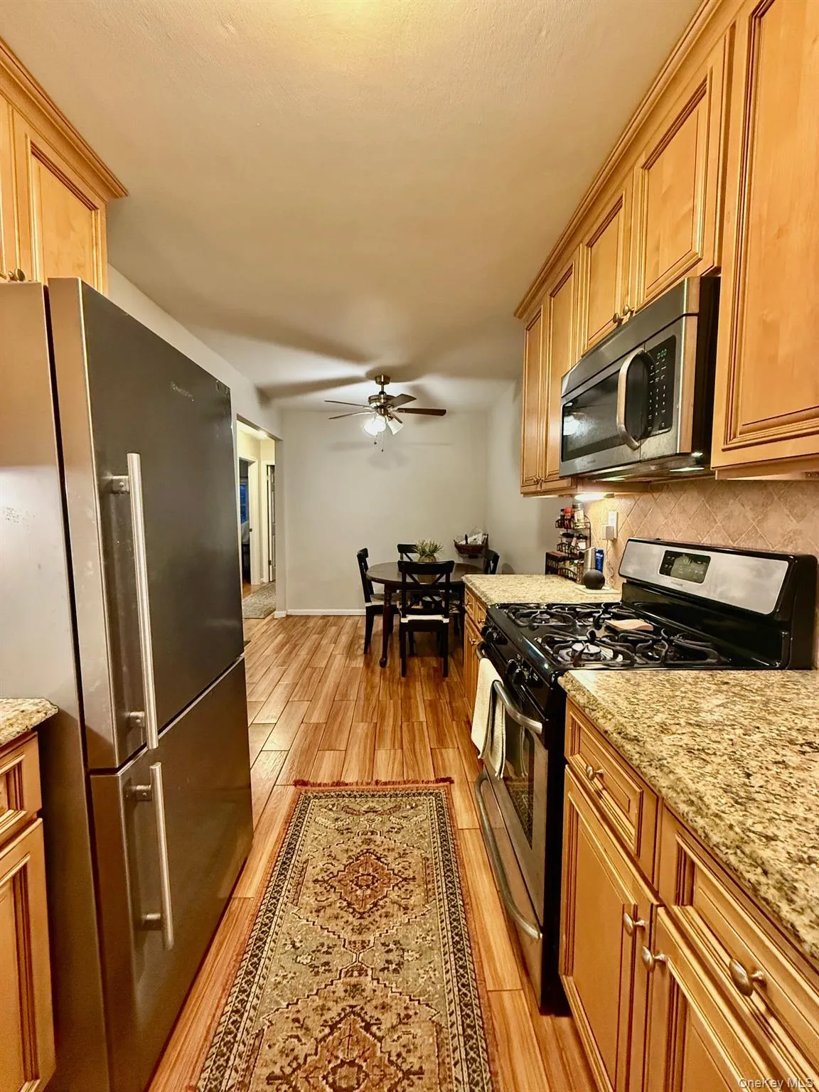 Kitchen featuring appliances with stainless steel finishes, light stone counters, light wood-style flooring, brown cabinetry, and a ceiling fan Kitchen featuring appliances with stainless steel finishes, light stone counters, light wood-style flooring, brown cabinetry, and a ceiling fan