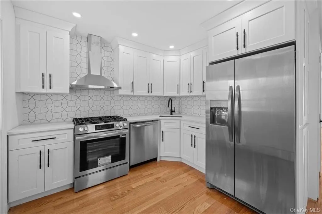 Kitchen featuring stainless steel appliances, white cabinets, wall chimney range hood, light wood finished floors, and recessed lighting Kitchen featuring stainless steel appliances, white cabinets, wall chimney range hood, light wood finished floors, and recessed lighting