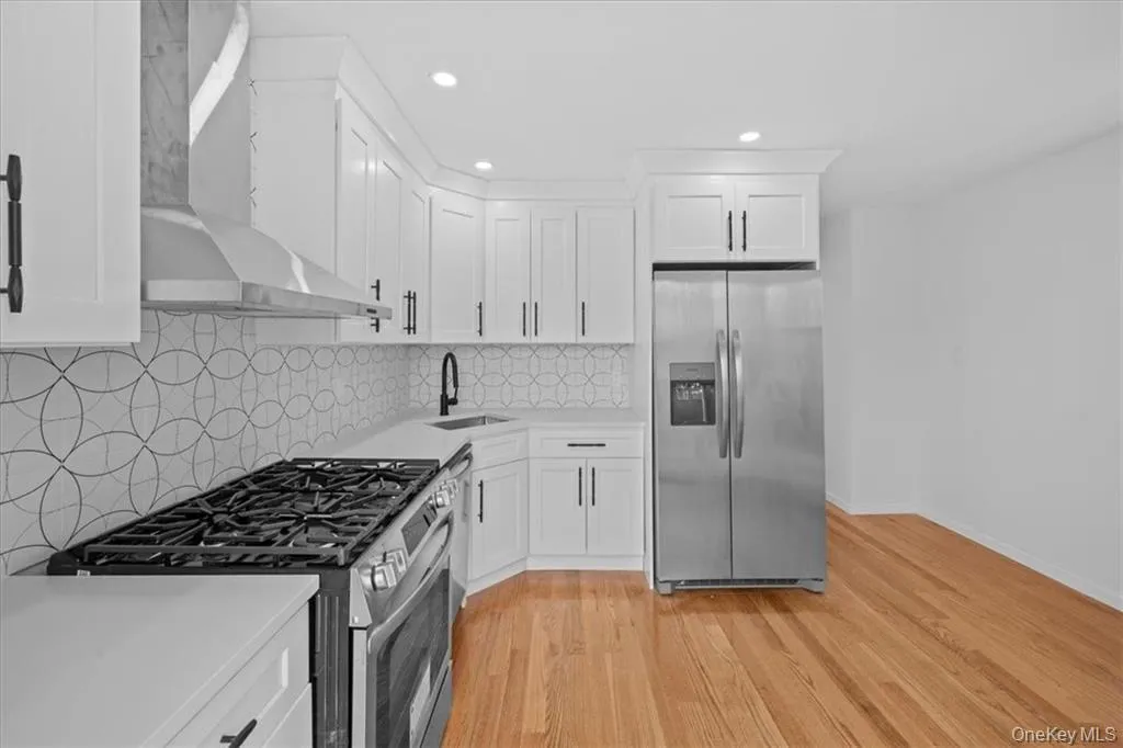Kitchen with appliances with stainless steel finishes, white cabinetry, wall chimney range hood, light wood-style flooring, and backsplash Kitchen with appliances with stainless steel finishes, white cabinetry, wall chimney range hood, light wood-style flooring, and backsplash