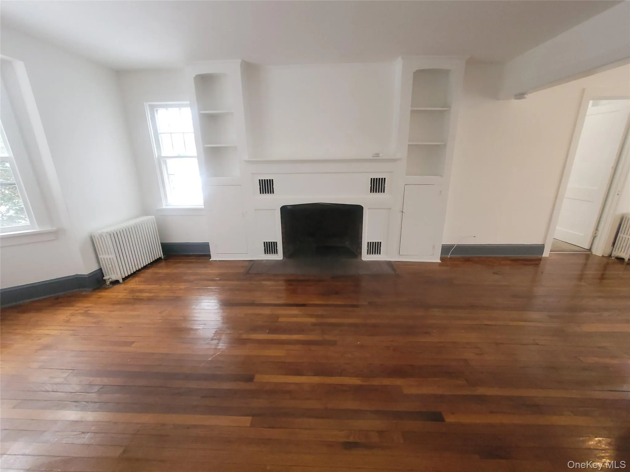 Unfurnished living room with radiator heating unit, a fireplace with flush hearth, built in features, and dark wood-style flooring Unfurnished living room with radiator heating unit, a fireplace with flush hearth, built in features, and dark wood-style flooring
