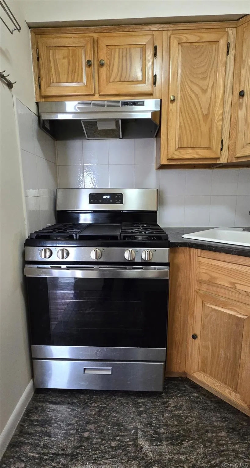 Kitchen with tasteful backsplash, gas stove, under cabinet range hood, dark countertops, and brown cabinetry Kitchen with tasteful backsplash, gas stove, under cabinet range hood, dark countertops, and brown cabinetry