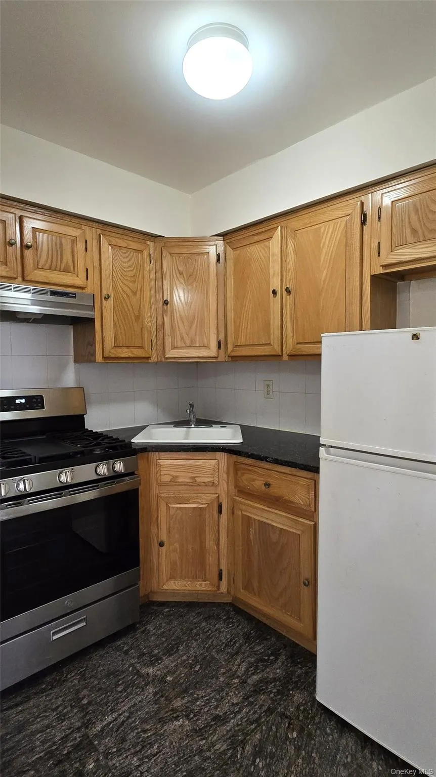 Kitchen with stainless steel range with gas stovetop, freestanding refrigerator, dark countertops, brown cabinets, and under cabinet range hood Kitchen with stainless steel range with gas stovetop, freestanding refrigerator, dark countertops, brown cabinets, and under cabinet range hood