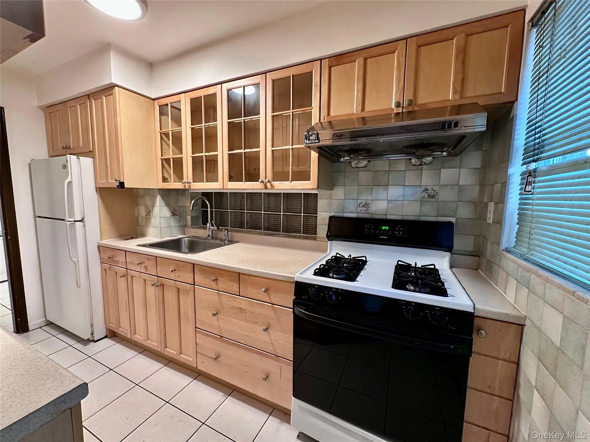 Kitchen with black gas range oven, light brown cabinets, exhaust hood, freestanding refrigerator, and glass insert cabinets Kitchen with black gas range oven, light brown cabinets, exhaust hood, freestanding refrigerator, and glass insert cabinets