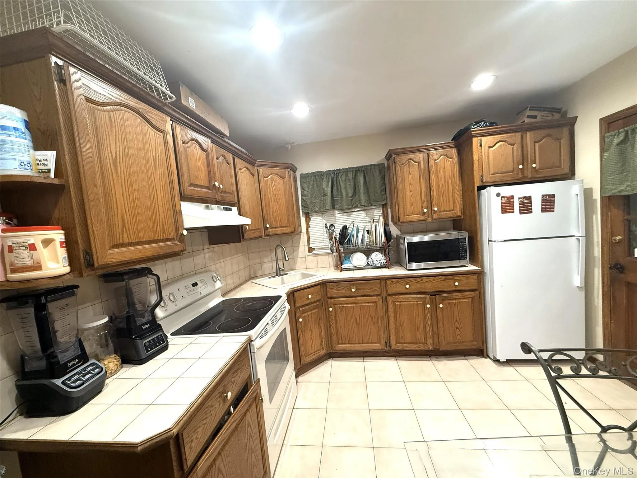 Kitchen with white appliances, brown cabinetry, backsplash, tile counters, and recessed lighting Kitchen with white appliances, brown cabinetry, backsplash, tile counters, and recessed lighting