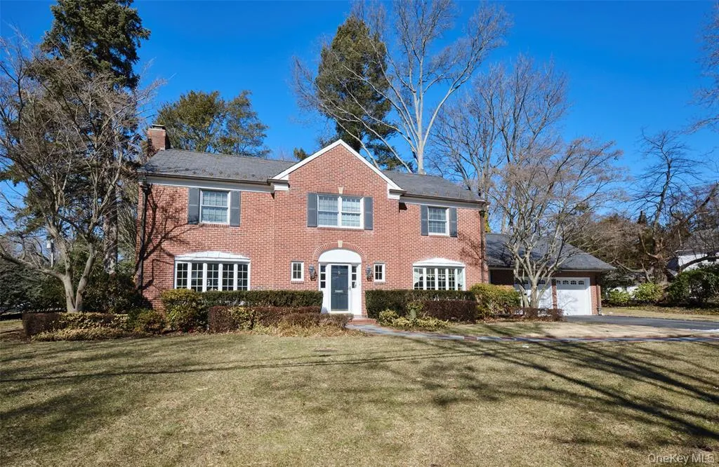 Colonial house with a front yard, an attached garage, a chimney, brick siding, and driveway Colonial house with a front yard, an attached garage, a chimney, brick siding, and driveway