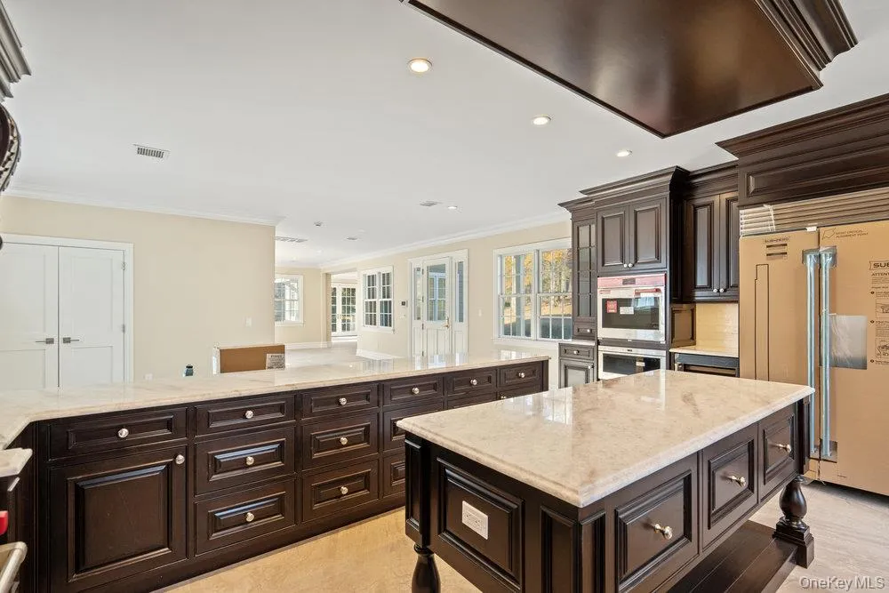 Kitchen with dark brown cabinetry, exhaust hood, paneled built in fridge, light stone countertops, and stainless steel double oven Kitchen with dark brown cabinetry, exhaust hood, paneled built in fridge, light stone countertops, and stainless steel double oven