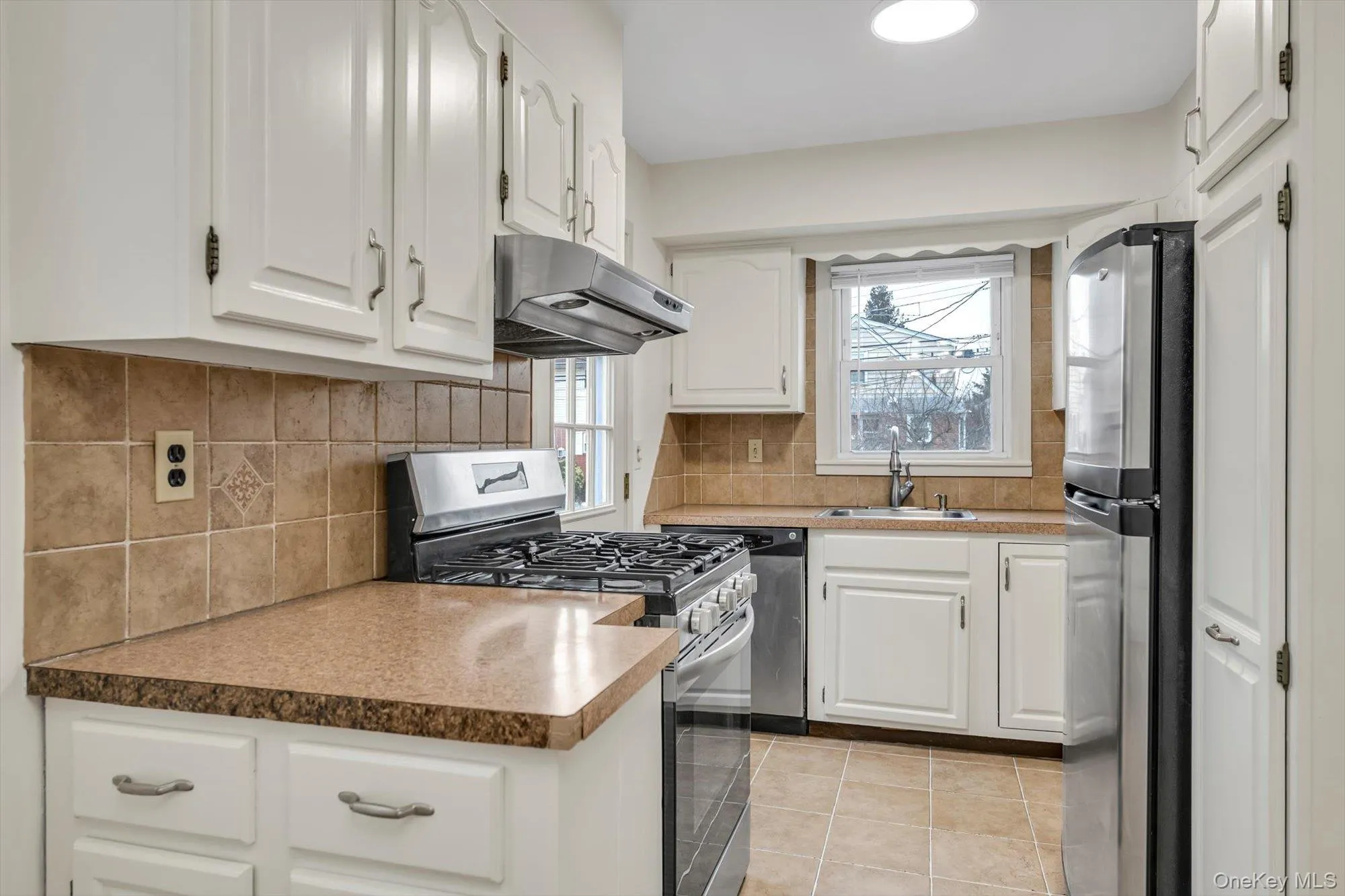 Kitchen with appliances with stainless steel finishes, white cabinetry, under cabinet range hood, light tile patterned flooring, and backsplash Kitchen with appliances with stainless steel finishes, white cabinetry, under cabinet range hood, light tile patterned flooring, and backsplash