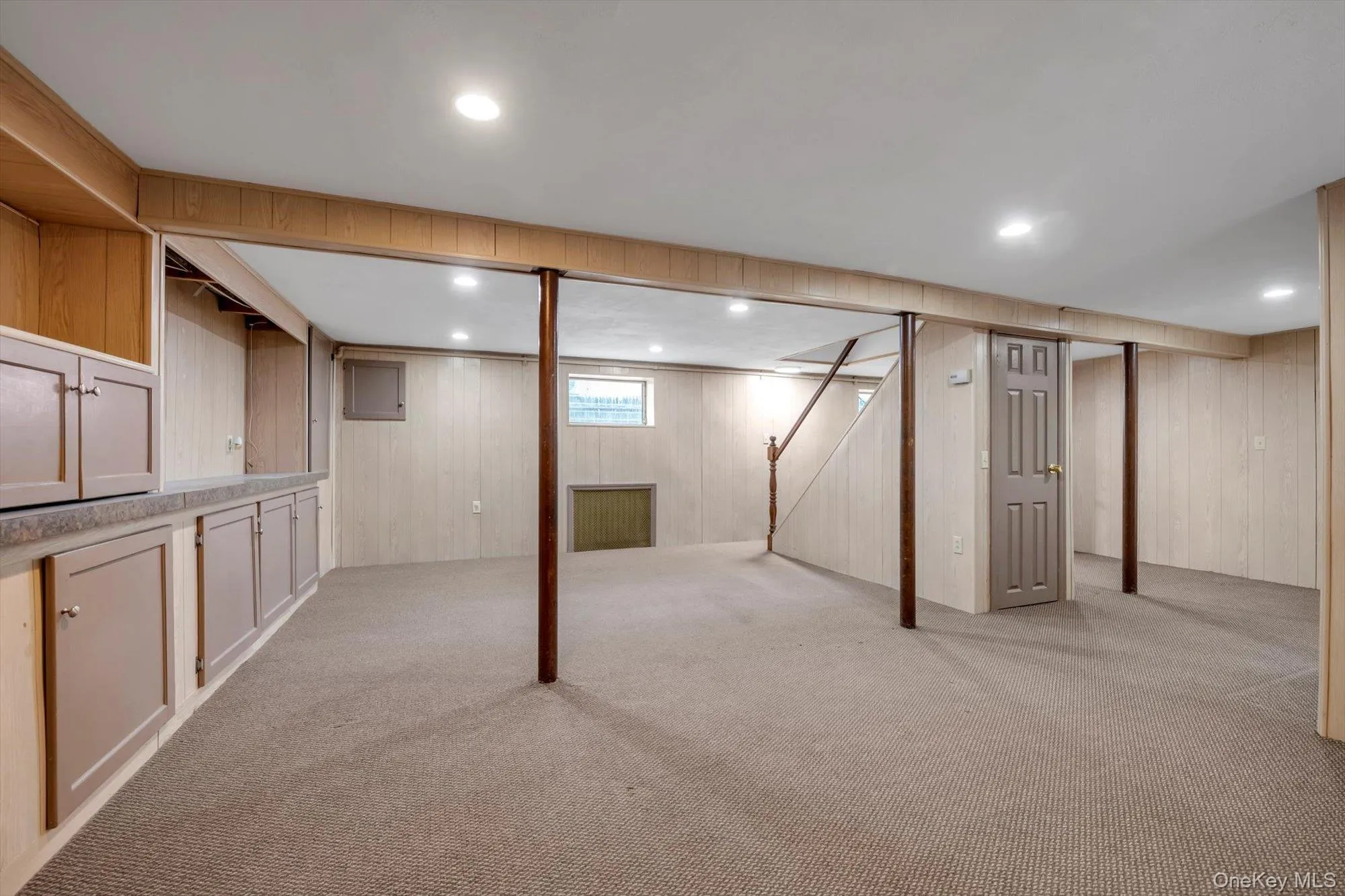 Basement with light carpet, wooden walls, radiator heating unit, electric panel, and stairs Basement with light carpet, wooden walls, radiator heating unit, electric panel, and stairs