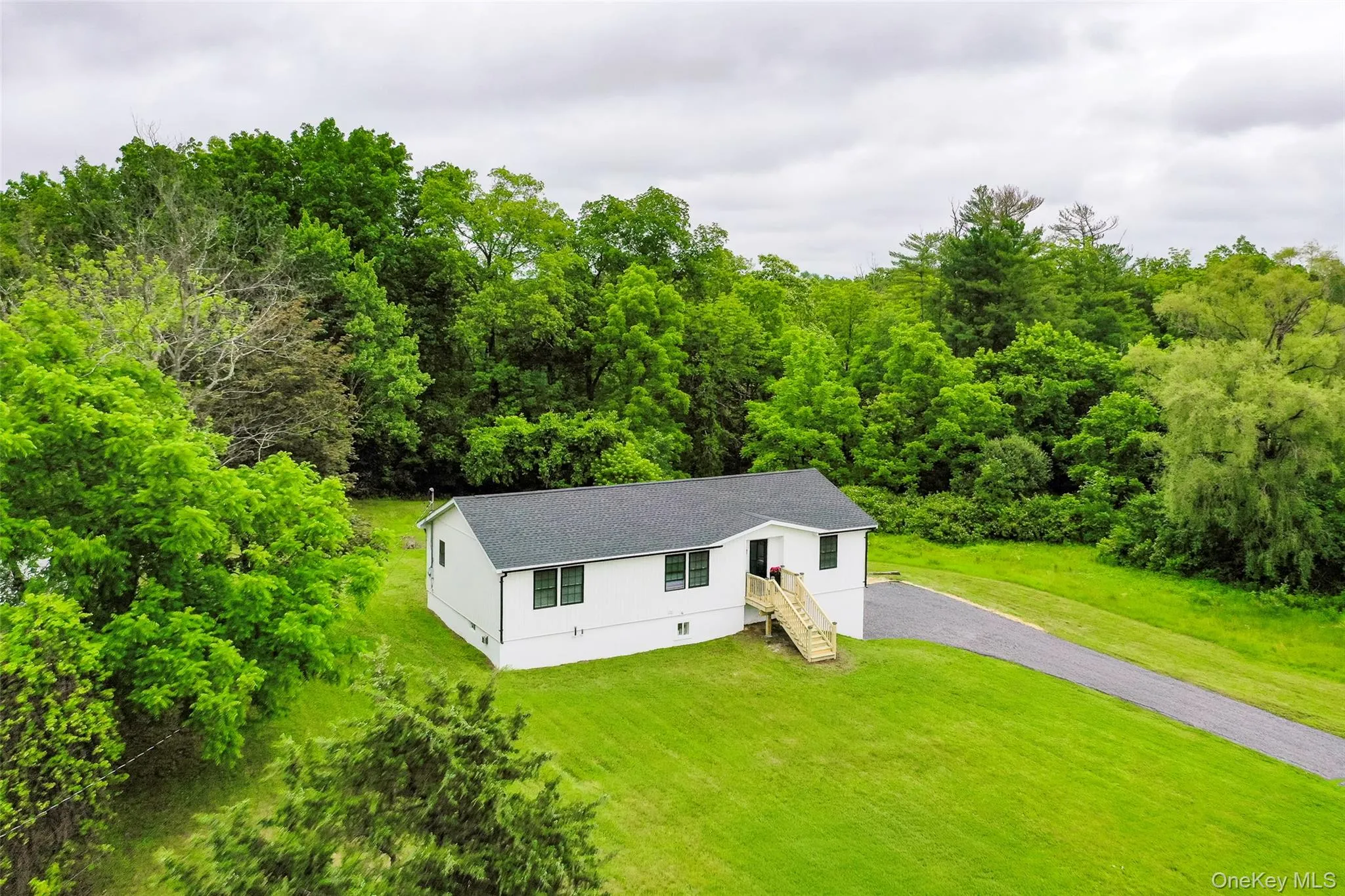 View of front facade featuring gravel driveway, a front lawn, a shingled roof, and a forest view View of front facade featuring gravel driveway, a front lawn, a shingled roof, and a forest view