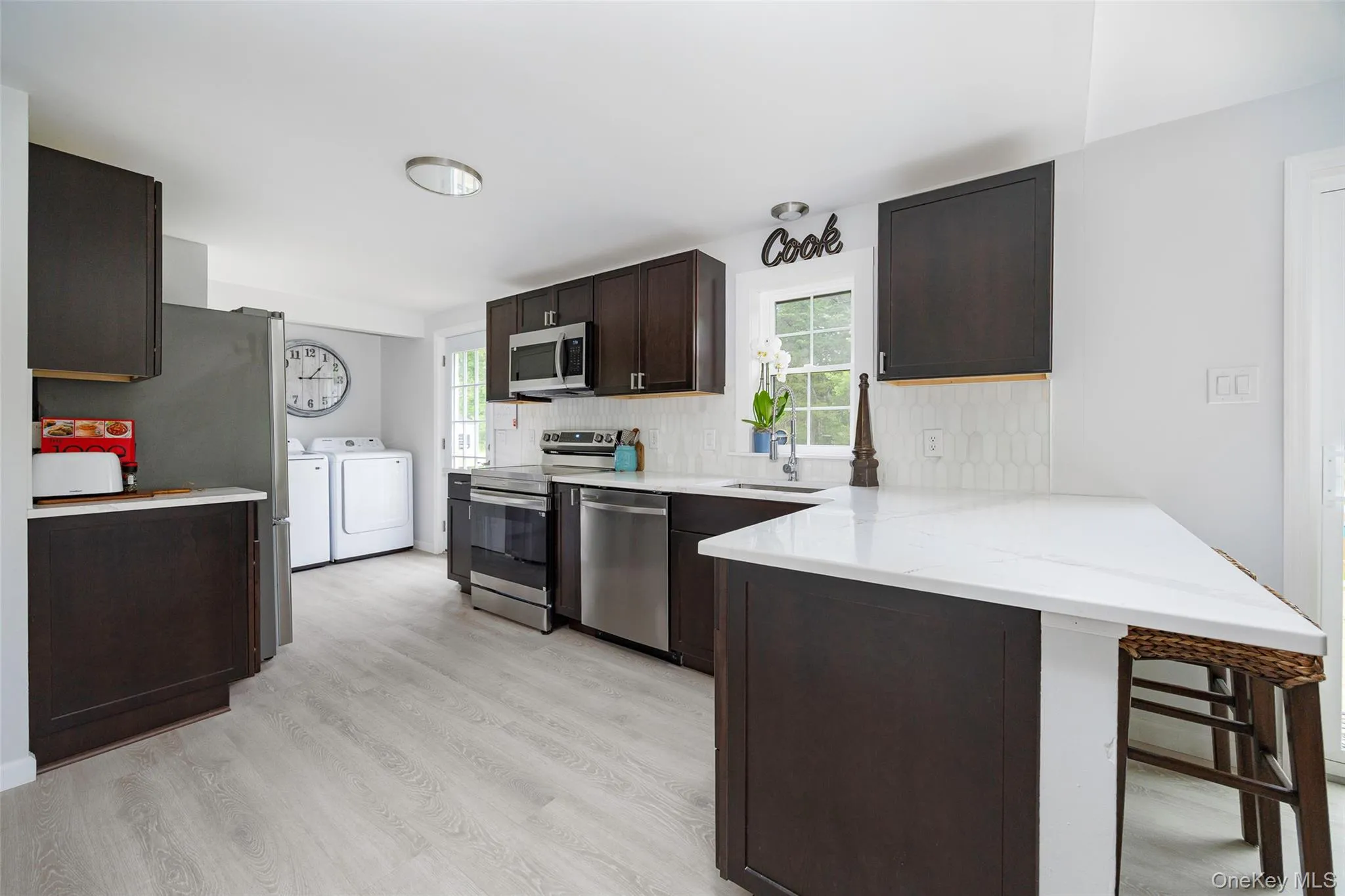 Kitchen featuring washer and dryer, appliances with stainless steel finishes, a peninsula, dark brown cabinetry, and tasteful backsplash Kitchen featuring washer and dryer, appliances with stainless steel finishes, a peninsula, dark brown cabinetry, and tasteful backsplash