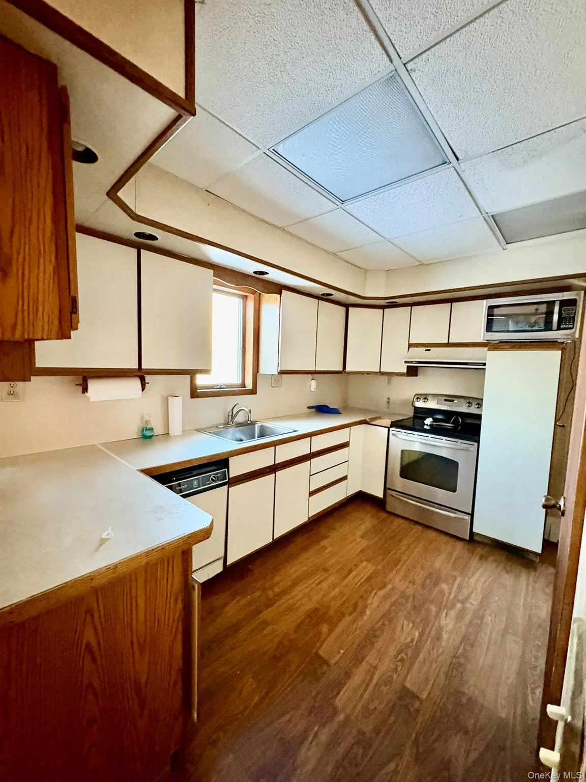 Kitchen featuring a drop ceiling, white cabinetry, brown cabinets, light countertops, and appliances with stainless steel finishes Kitchen featuring a drop ceiling, white cabinetry, brown cabinets, light countertops, and appliances with stainless steel finishes