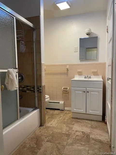 Bathroom featuring vanity, a baseboard heating unit, shower / bath combination with glass door, a wainscoted wall, and tile walls Bathroom featuring vanity, a baseboard heating unit, shower / bath combination with glass door, a wainscoted wall, and tile walls