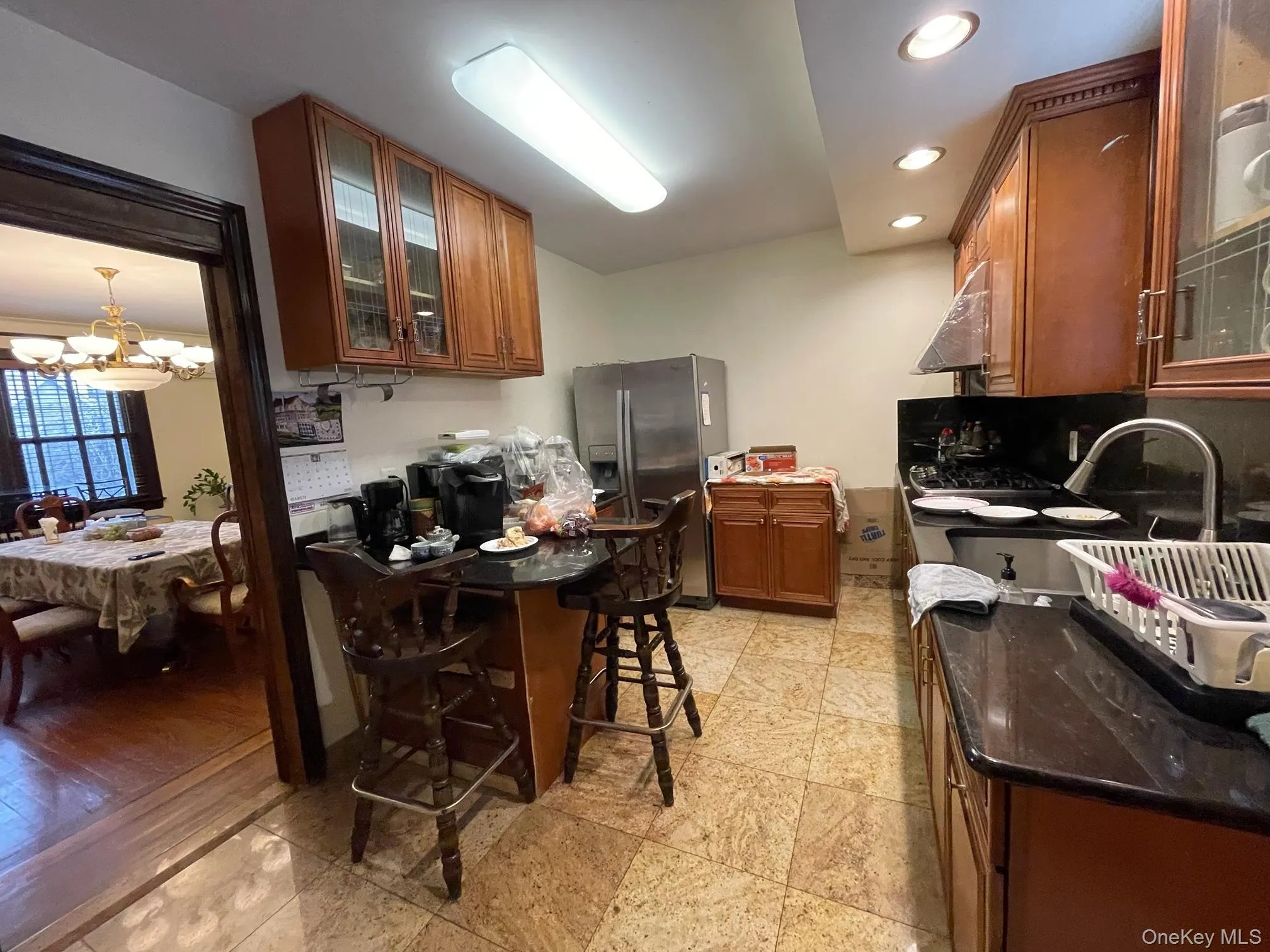 Kitchen featuring glass insert cabinets, brown cabinetry, stainless steel fridge with ice dispenser, a chandelier, and recessed lighting Kitchen featuring glass insert cabinets, brown cabinetry, stainless steel fridge with ice dispenser, a chandelier, and recessed lighting