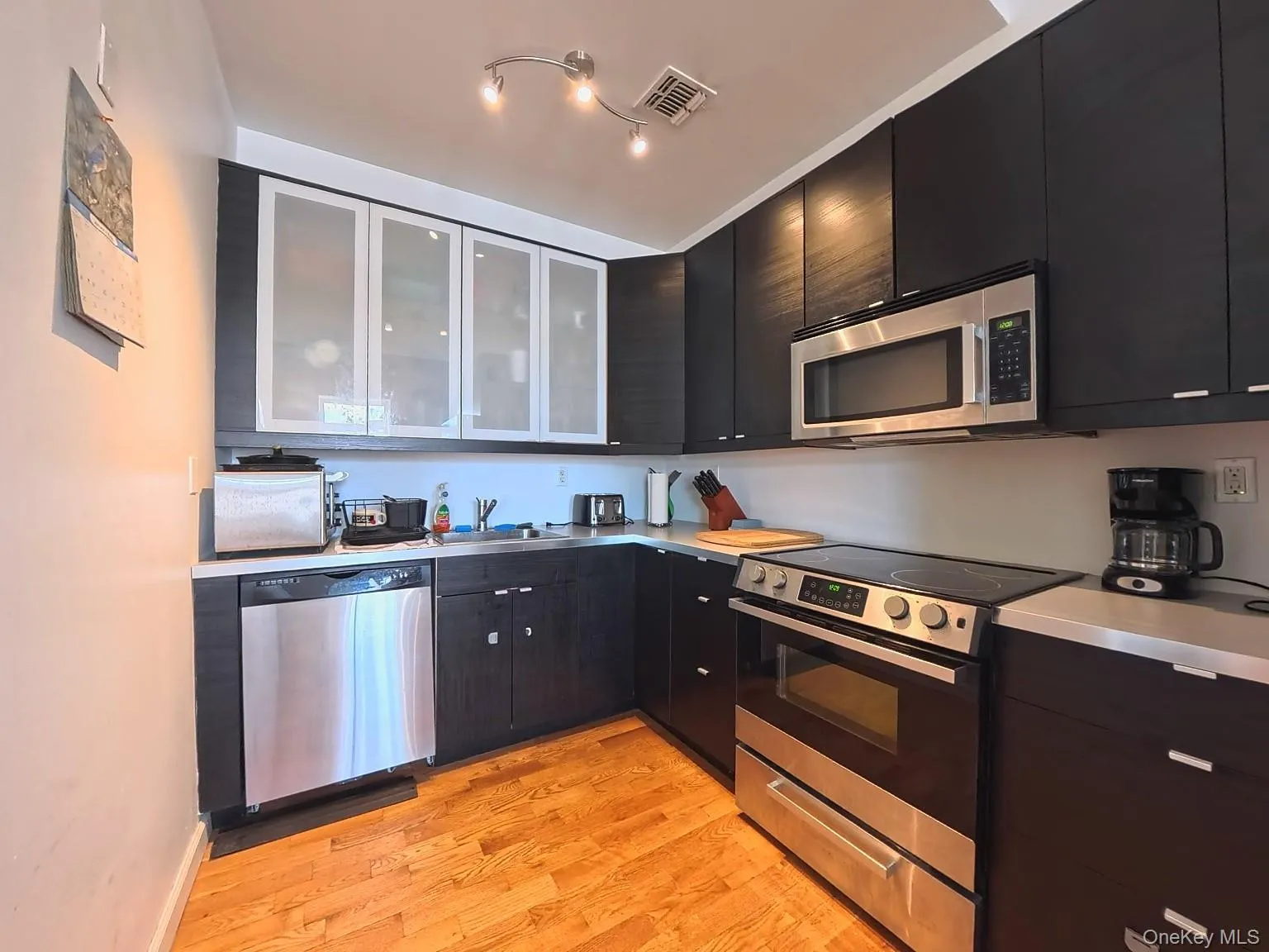 Kitchen with appliances with stainless steel finishes, dark cabinets, light countertops, light wood-style floors, and glass insert cabinets Kitchen with appliances with stainless steel finishes, dark cabinets, light countertops, light wood-style floors, and glass insert cabinets