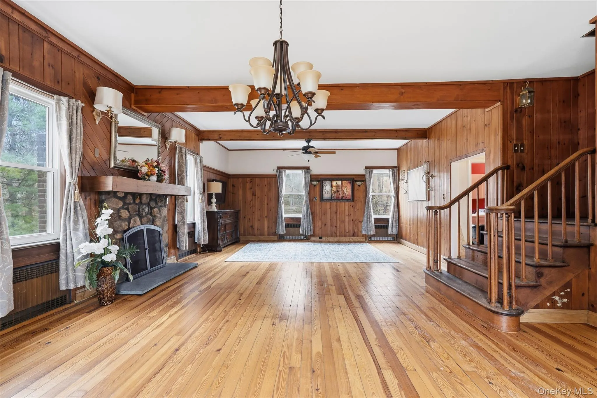 Unfurnished living room with wooden walls, a stone fireplace, light wood finished floors, stairs, and beamed ceiling Unfurnished living room with wooden walls, a stone fireplace, light wood finished floors, stairs, and beamed ceiling