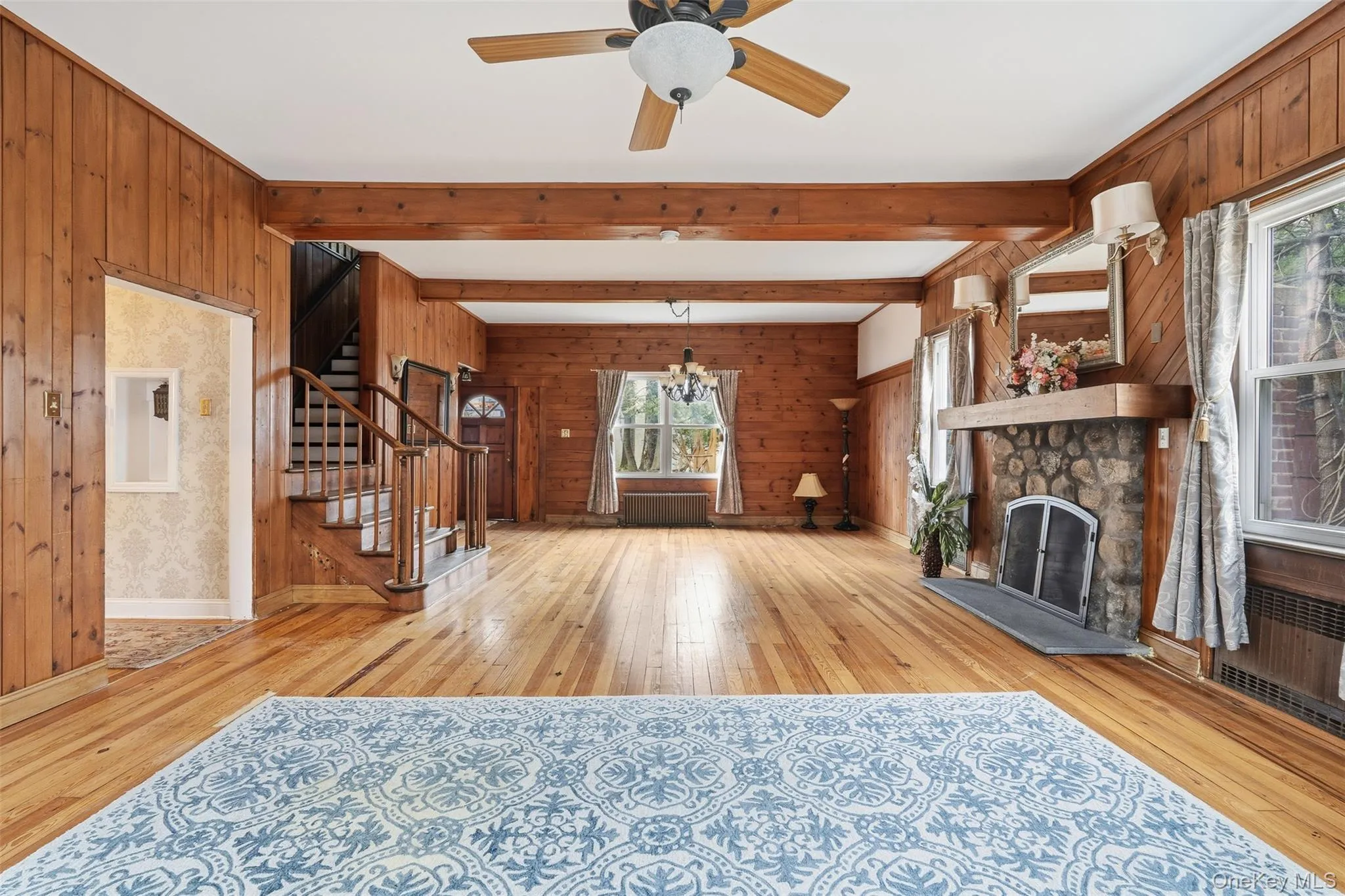 Unfurnished living room with wood walls, light wood-style flooring, a chandelier, beamed ceiling, and stairs Unfurnished living room with wood walls, light wood-style flooring, a chandelier, beamed ceiling, and stairs