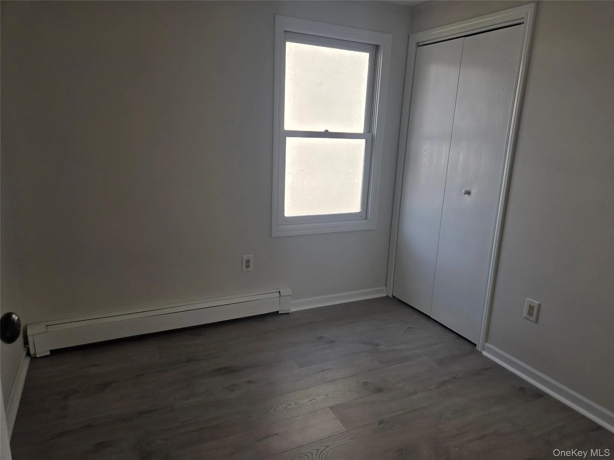 Unfurnished bedroom with baseboard heating, dark wood-style flooring, and a closet Unfurnished bedroom with baseboard heating, dark wood-style flooring, and a closet