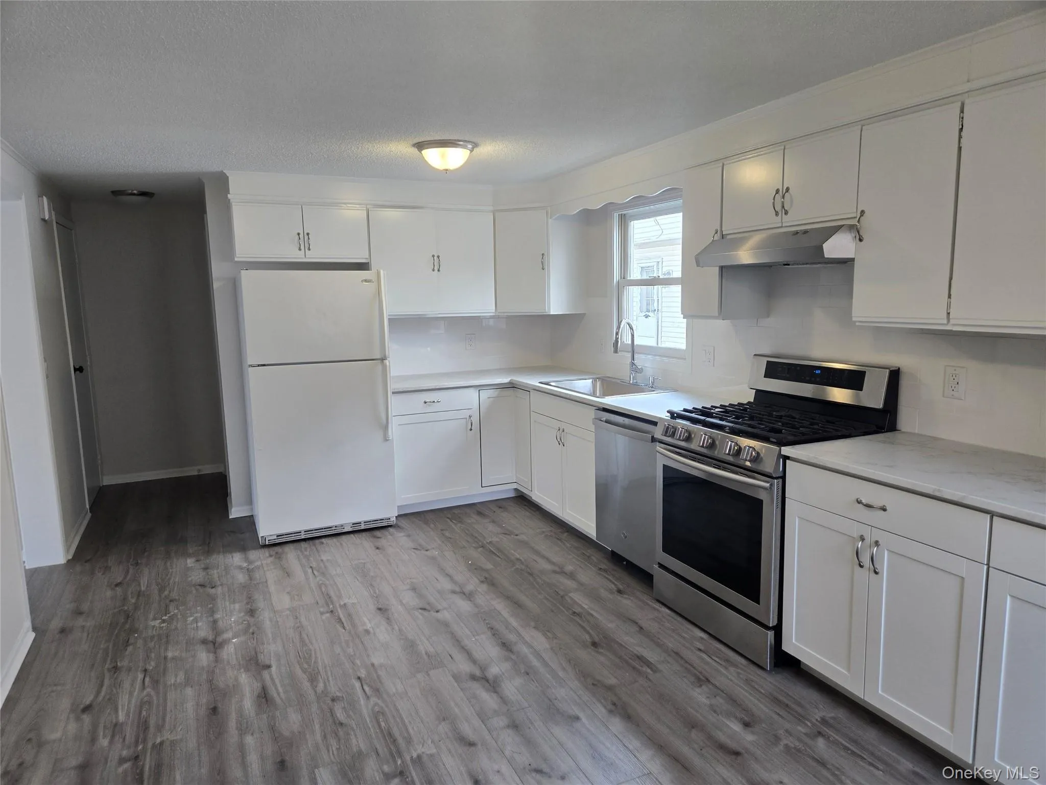 Kitchen with stainless steel appliances, white cabinets, under cabinet range hood, light wood-type flooring, and a textured ceiling Kitchen with stainless steel appliances, white cabinets, under cabinet range hood, light wood-type flooring, and a textured ceiling