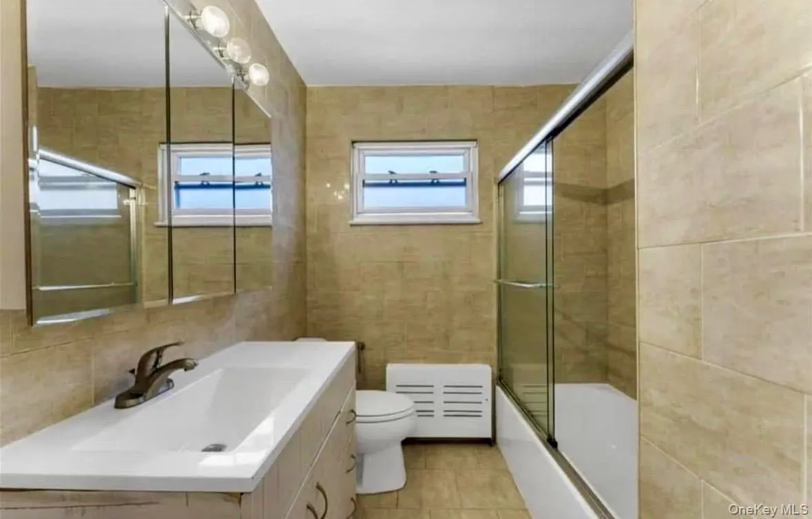 Full bathroom with tile walls, vanity, bath / shower combo with glass door, and light tile patterned floors Full bathroom with tile walls, vanity, bath / shower combo with glass door, and light tile patterned floors