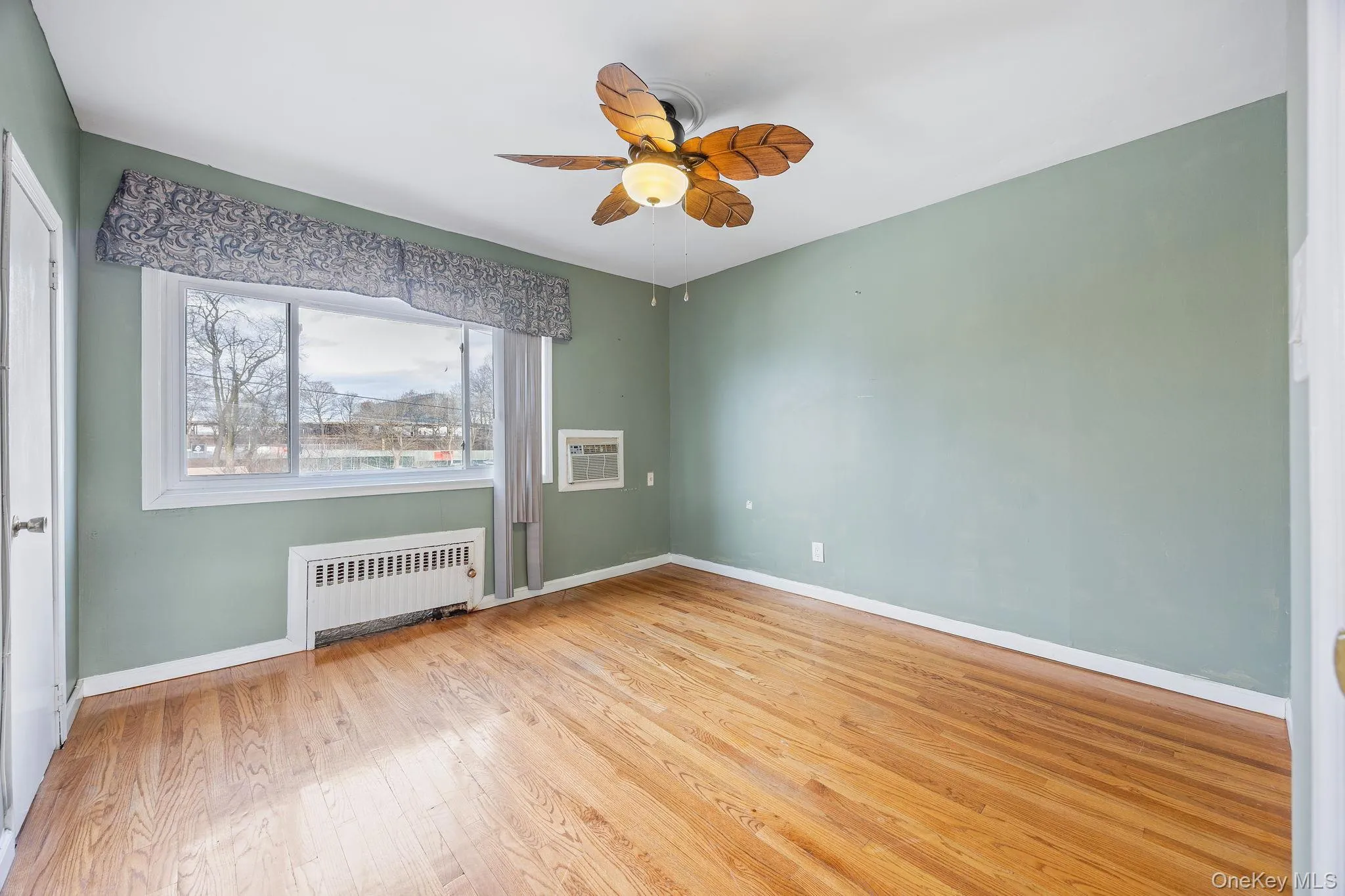 Unfurnished room with radiator heating unit, light wood-style flooring, ceiling fan, and a wall unit AC Unfurnished room with radiator heating unit, light wood-style flooring, ceiling fan, and a wall unit AC