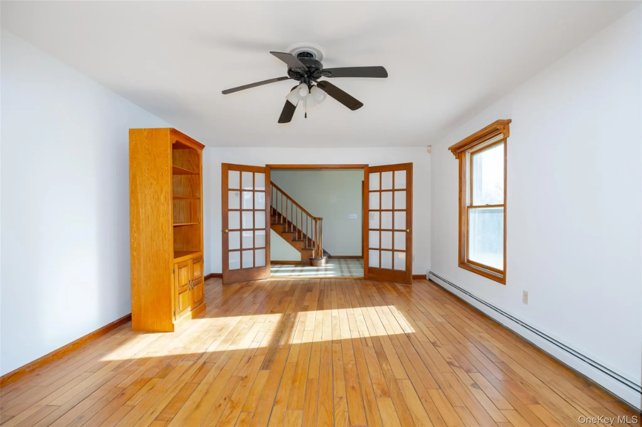 Unfurnished living room featuring a baseboard radiator, french doors, stairs, light wood floors, and a ceiling fan Unfurnished living room featuring a baseboard radiator, french doors, stairs, light wood floors, and a ceiling fan