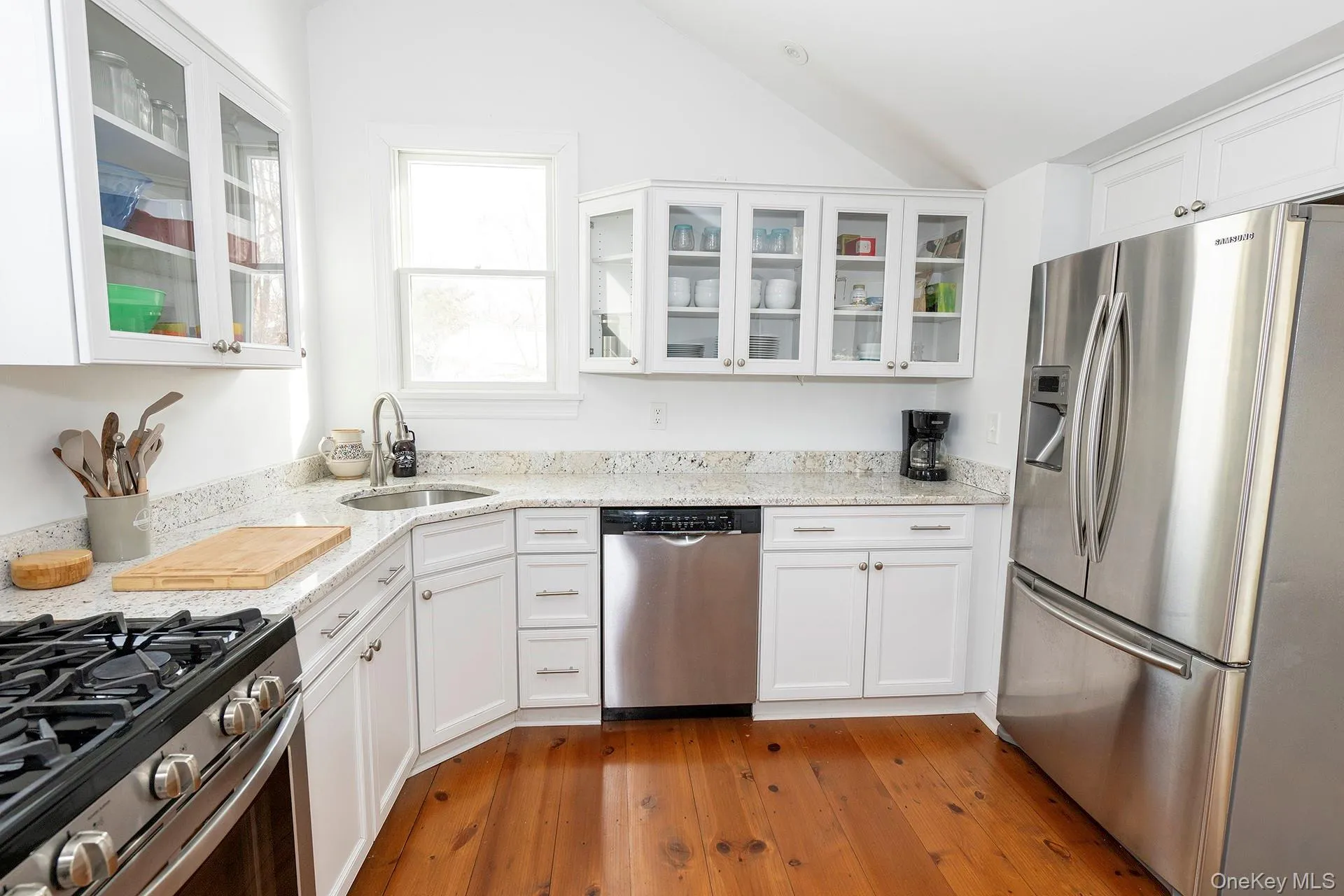 Kitchen with appliances with stainless steel finishes, glass insert cabinets, white cabinetry, light stone countertops, and dark wood-style floors Kitchen with appliances with stainless steel finishes, glass insert cabinets, white cabinetry, light stone countertops, and dark wood-style floors