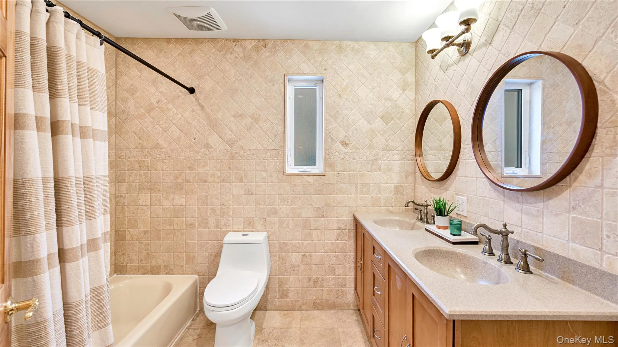 Bathroom with tile walls, double vanity, shower / bath combo with shower curtain, and light tile patterned floors Bathroom with tile walls, double vanity, shower / bath combo with shower curtain, and light tile patterned floors