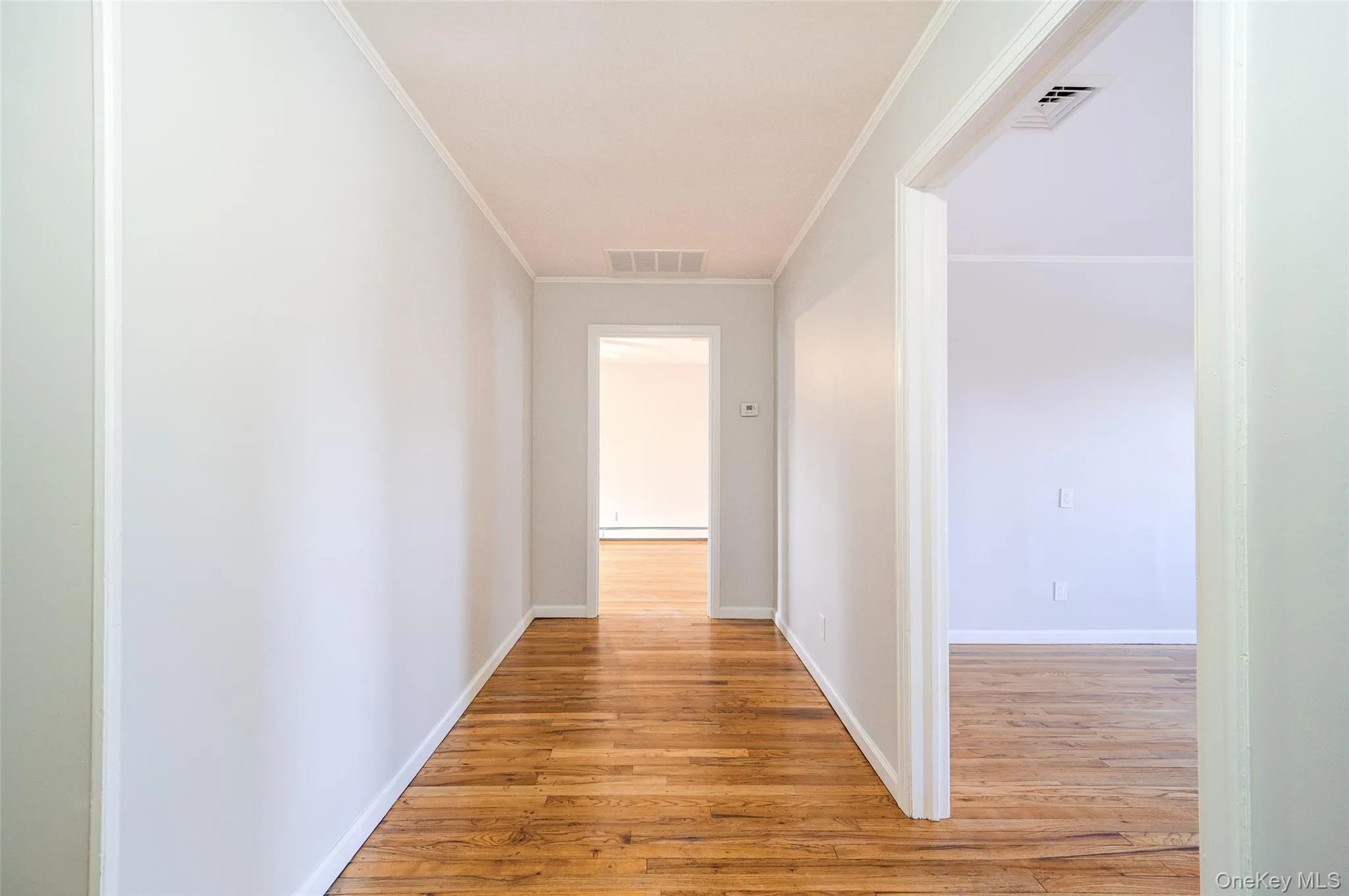 Hallway to living room (on right) and kitchen (ahead) featuring light wood finished floors, ornamental molding, and a baseboard heating unit Hallway to living room (on right) and kitchen (ahead) featuring light wood finished floors, ornamental molding, and a baseboard heating unit