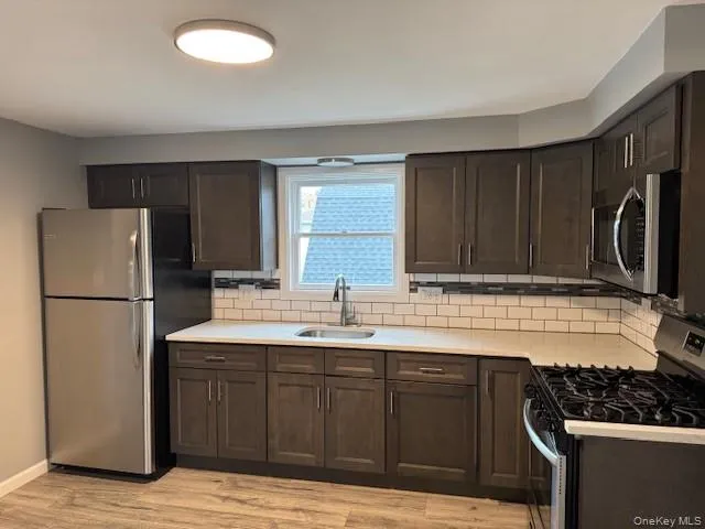 Kitchen featuring dark brown cabinetry, appliances with stainless steel finishes, backsplash, light wood-style flooring, and light stone countertops Kitchen featuring dark brown cabinetry, appliances with stainless steel finishes, backsplash, light wood-style flooring, and light stone countertops