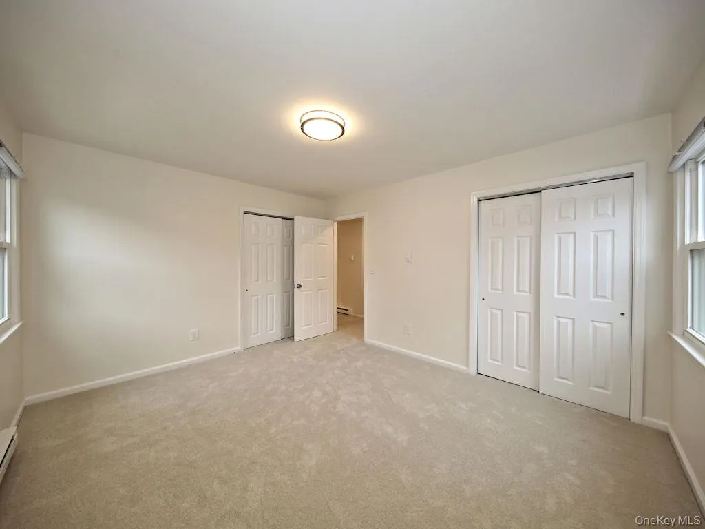 Unfurnished bedroom featuring two closets, light carpet, and a baseboard heating unit Unfurnished bedroom featuring two closets, light carpet, and a baseboard heating unit