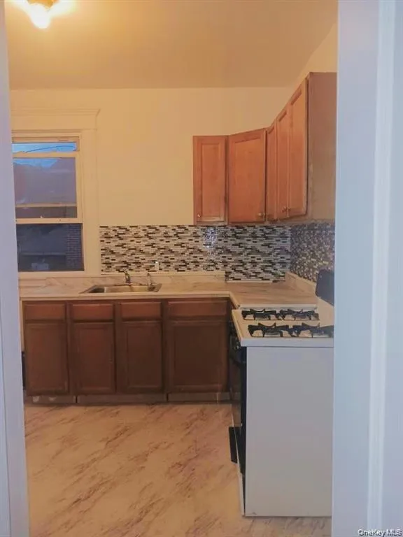 Kitchen featuring light countertops, white range with gas stovetop, backsplash, and brown cabinets Kitchen featuring light countertops, white range with gas stovetop, backsplash, and brown cabinets