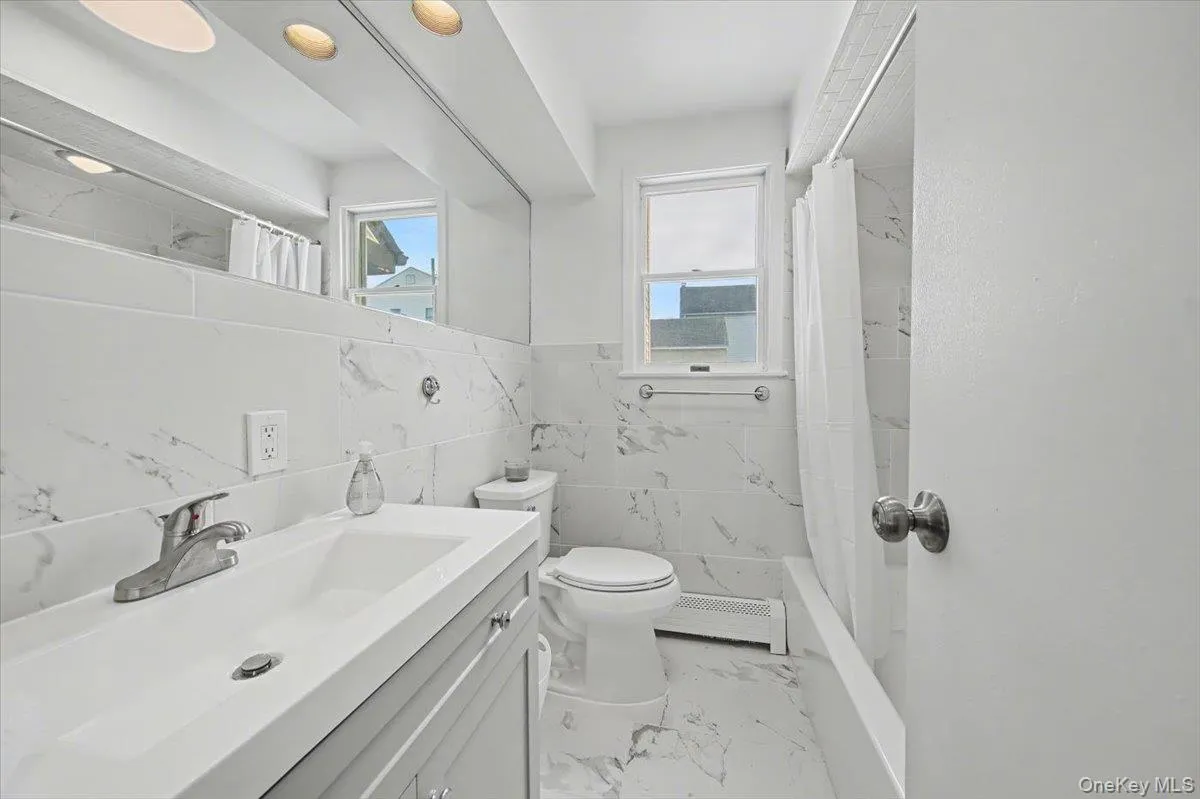 Full bath featuring tile walls, vanity, light marble finish flooring, a wainscoted wall, and baseboard heating Full bath featuring tile walls, vanity, light marble finish flooring, a wainscoted wall, and baseboard heating