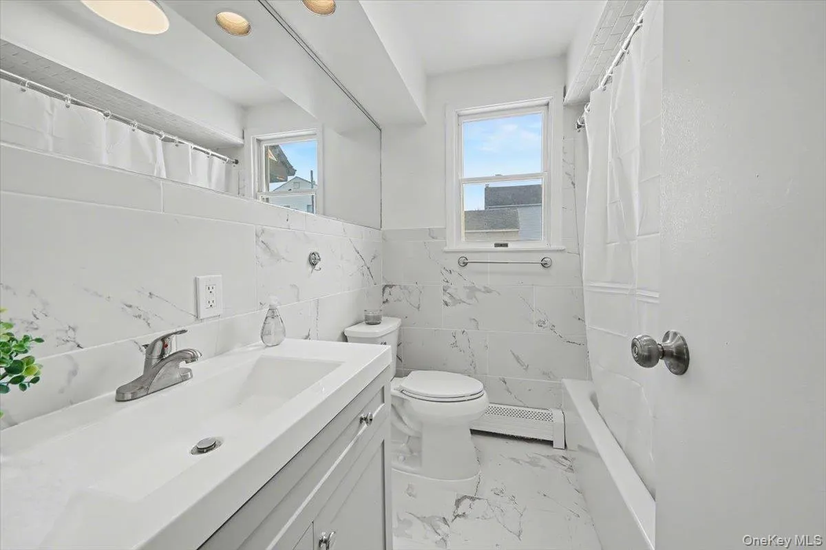 Full bathroom featuring tile walls, vanity, light marble finish flooring, baseboard heating, and wainscoting Full bathroom featuring tile walls, vanity, light marble finish flooring, baseboard heating, and wainscoting