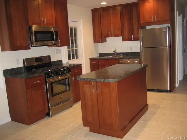 Kitchen with appliances with stainless steel finishes, a kitchen island, light tile patterned flooring, recessed lighting, and brown cabinetry Kitchen with appliances with stainless steel finishes, a kitchen island, light tile patterned flooring, recessed lighting, and brown cabinetry