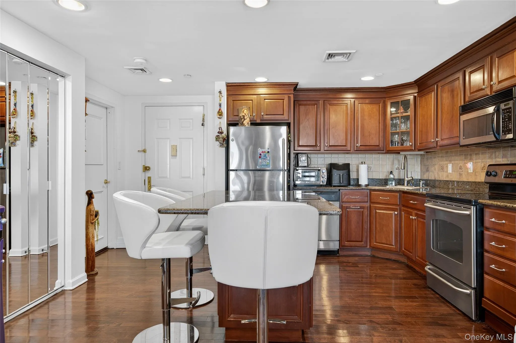 Kitchen with stainless steel appliances, brown cabinetry, a kitchen bar, glass insert cabinets, and dark stone counters Kitchen with stainless steel appliances, brown cabinetry, a kitchen bar, glass insert cabinets, and dark stone counters