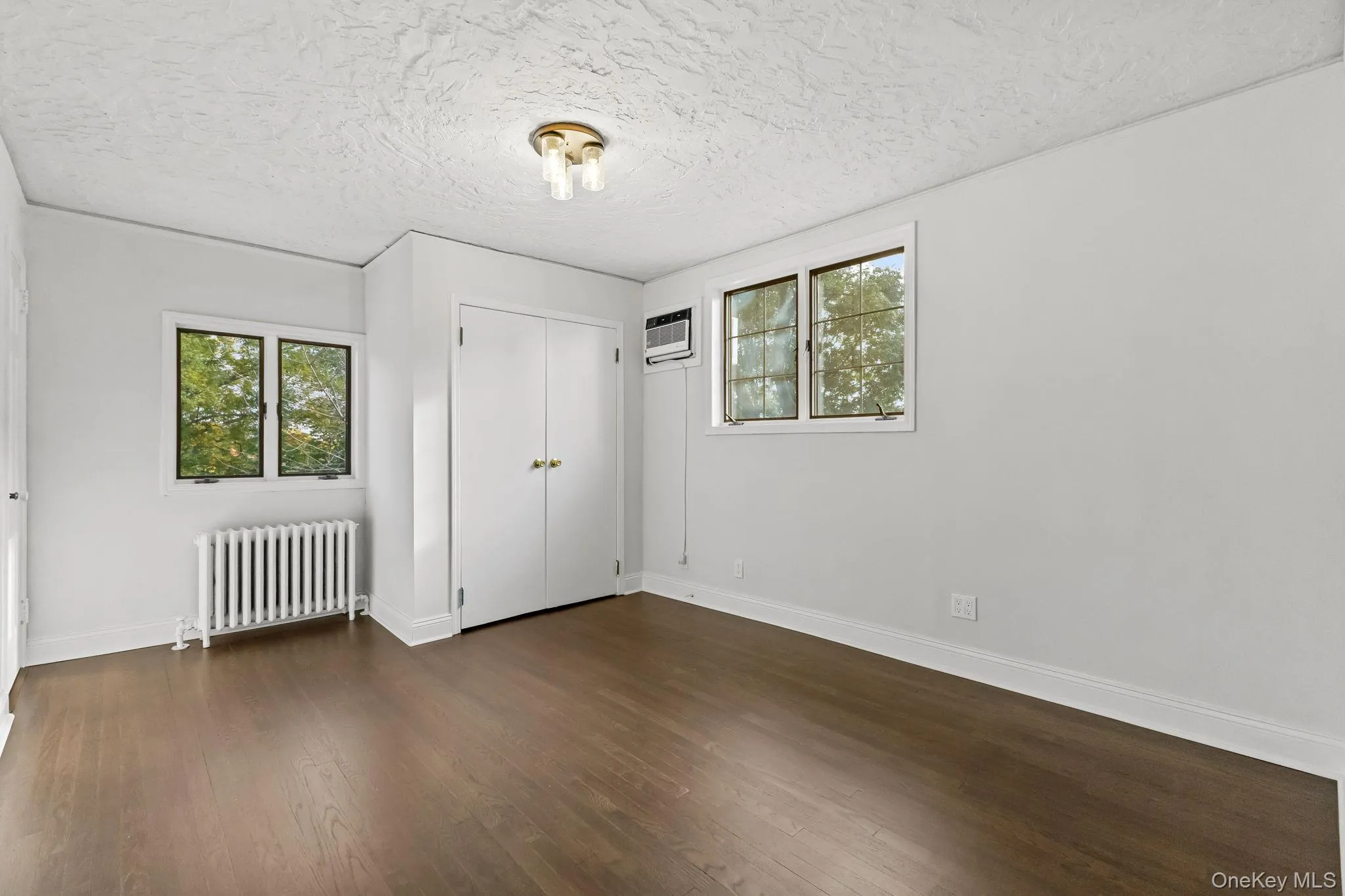 Unfurnished bedroom with radiator heating unit, dark wood finished floors, a closet, a textured ceiling, and a wall mounted AC Unfurnished bedroom with radiator heating unit, dark wood finished floors, a closet, a textured ceiling, and a wall mounted AC