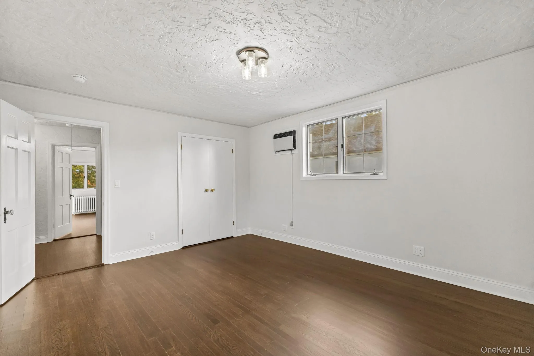 Unfurnished bedroom featuring dark wood-style flooring, a closet, a textured ceiling, radiator, and a wall mounted air conditioner Unfurnished bedroom featuring dark wood-style flooring, a closet, a textured ceiling, radiator, and a wall mounted air conditioner