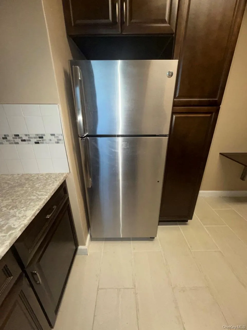 Kitchen featuring dark brown cabinets, freestanding refrigerator, light stone countertops, and backsplash Kitchen featuring dark brown cabinets, freestanding refrigerator, light stone countertops, and backsplash