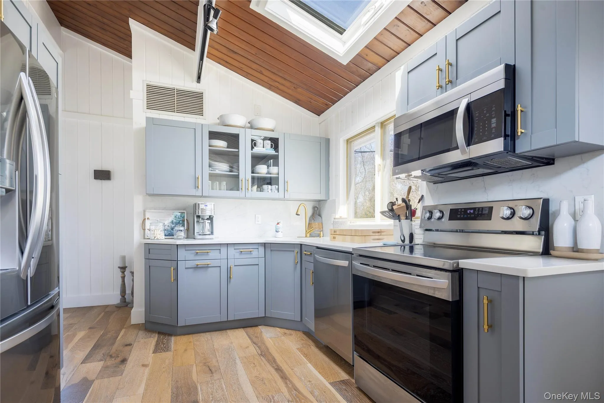 Kitchen with stainless steel appliances, gray cabinets, a skylight, glass insert cabinets, and light wood-type flooring Kitchen with stainless steel appliances, gray cabinets, a skylight, glass insert cabinets, and light wood-type flooring