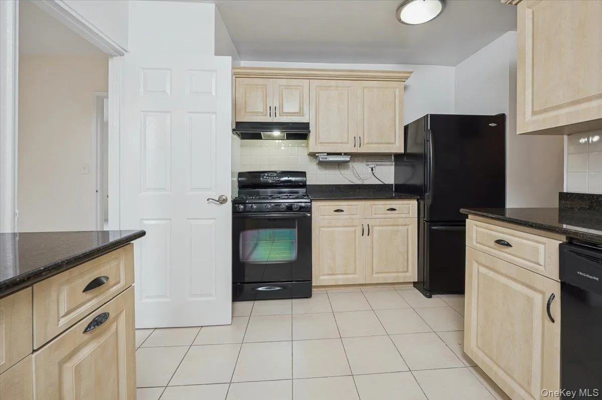 Kitchen with light brown cabinets, black appliances, dark stone countertops, light tile patterned flooring, and under cabinet range hood Kitchen with light brown cabinets, black appliances, dark stone countertops, light tile patterned flooring, and under cabinet range hood