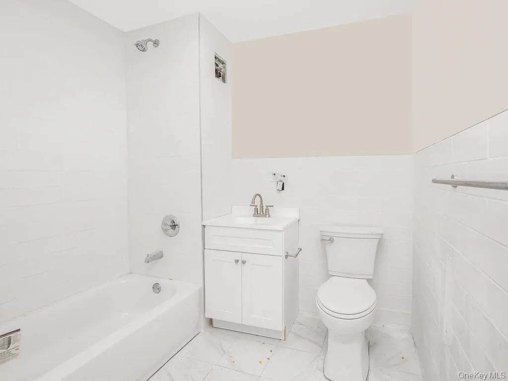 Bathroom featuring bathing tub / shower combination, vanity, tile walls, light marble finish floors, and a wainscoted wall Bathroom featuring bathing tub / shower combination, vanity, tile walls, light marble finish floors, and a wainscoted wall