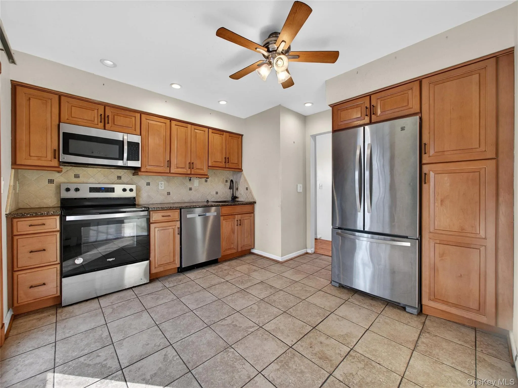 Kitchen featuring stainless steel appliances, ceiling fan, brown cabinets, backsplash, and light tile patterned flooring Kitchen featuring stainless steel appliances, ceiling fan, brown cabinets, backsplash, and light tile patterned flooring