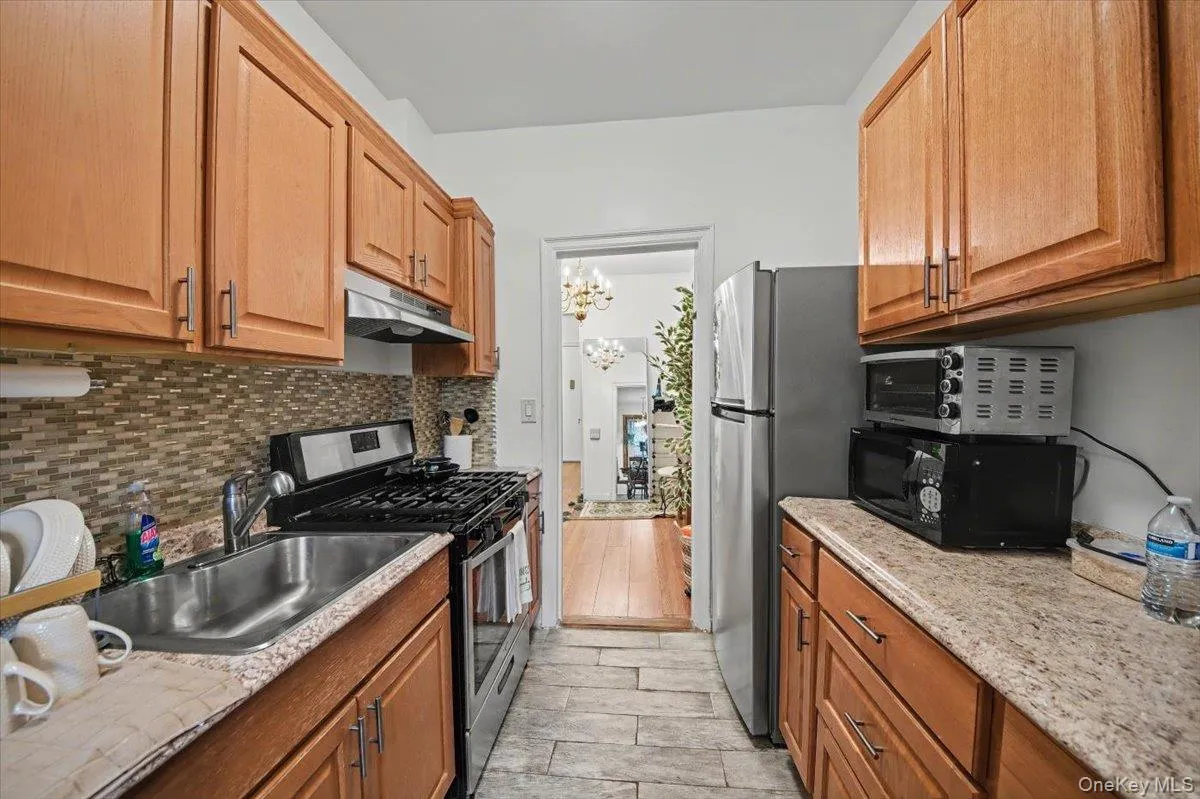 Kitchen featuring stainless steel appliances, a chandelier, brown cabinetry, under cabinet range hood, and wood finish floors Kitchen featuring stainless steel appliances, a chandelier, brown cabinetry, under cabinet range hood, and wood finish floors