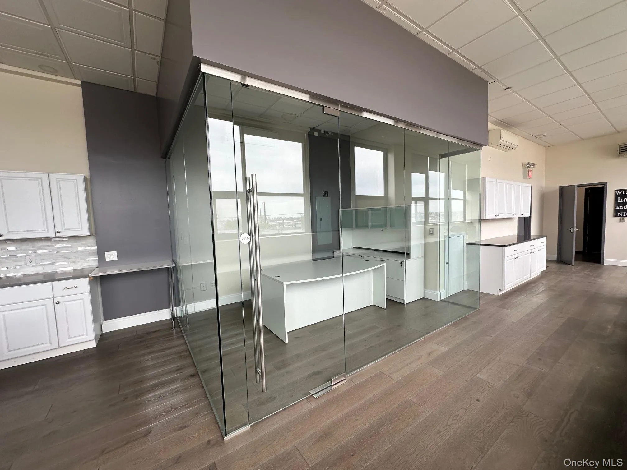 Kitchen featuring white cabinets, tasteful backsplash, dark wood-style floors, an AC wall unit, and a drop ceiling Kitchen featuring white cabinets, tasteful backsplash, dark wood-style floors, an AC wall unit, and a drop ceiling