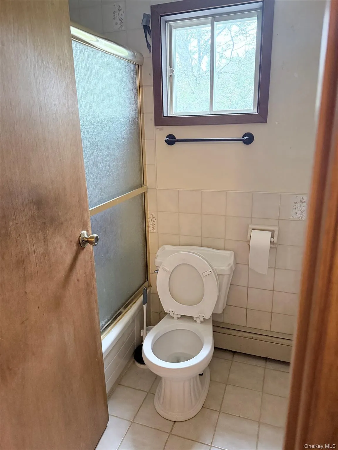 Full bath with light tile patterned floors, bath / shower combo with glass door, baseboard heating, and tile walls Full bath with light tile patterned floors, bath / shower combo with glass door, baseboard heating, and tile walls