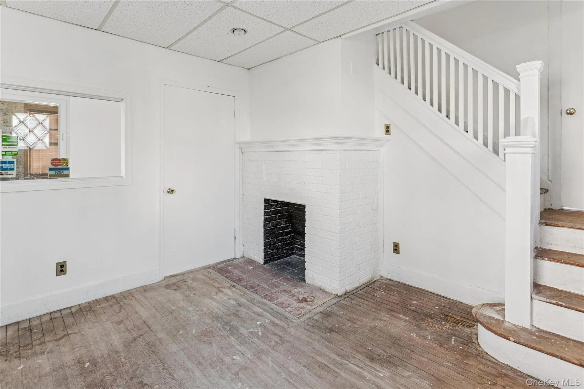 Unfurnished living room featuring stairway, a drop ceiling, wood-type flooring, and a brick fireplace Unfurnished living room featuring stairway, a drop ceiling, wood-type flooring, and a brick fireplace
